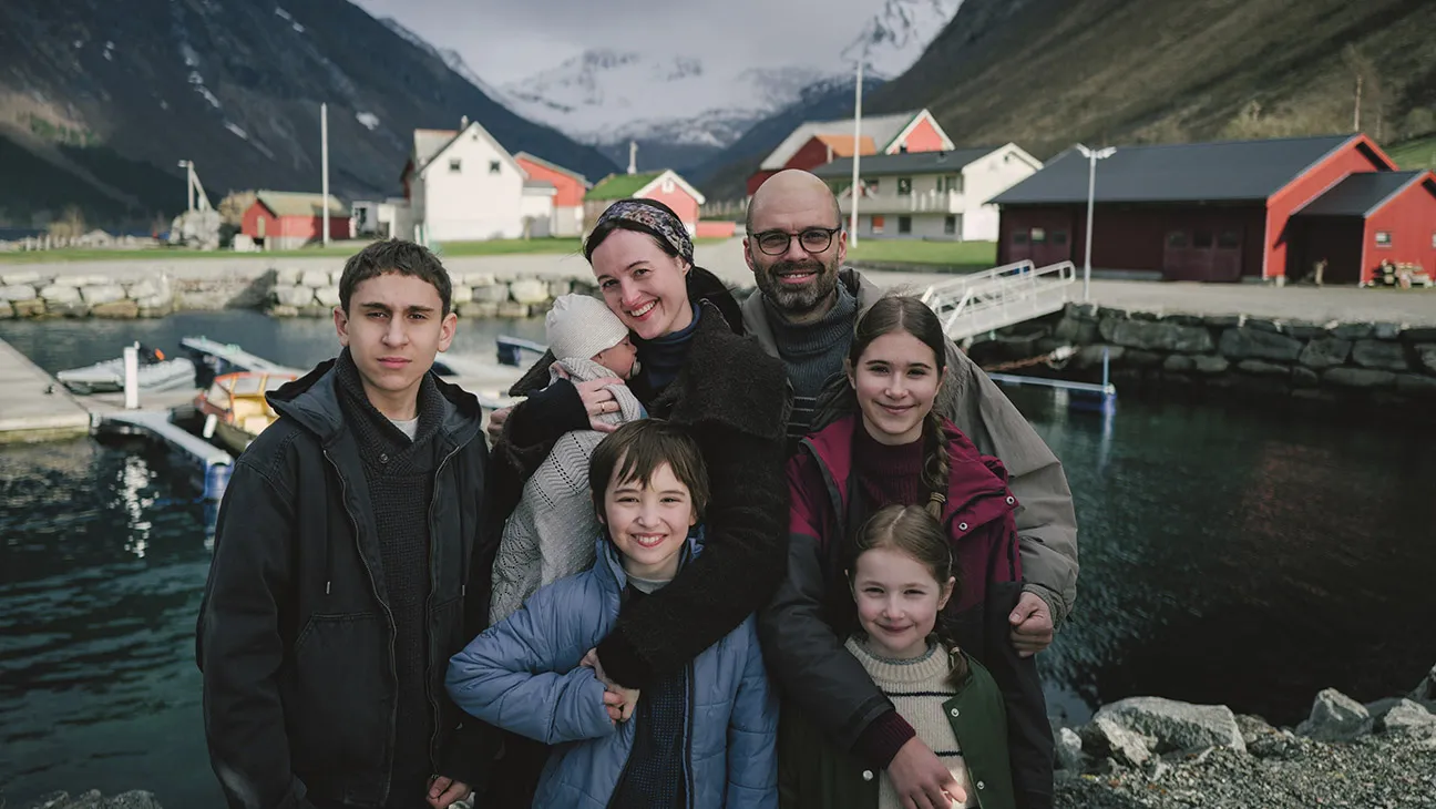 A family portrait still from 'Fjord': Sebastian Stan and Renate Reinsve with their five children, posed by a fjord with a Norwegian village and snow-capped mountains behind them.